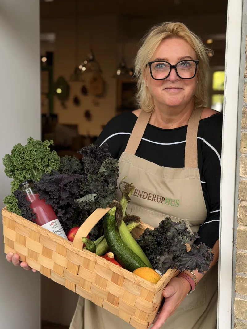 Mette i BrenderupHus-forklæde i døren med en kurv fuld af grønkål, squash, tomater og saft.